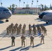 A formal group photograph of U.S. Air Force personnel standing in formation on a flight line tarmac at March Field Air Reserve Base. Two C-130 Hercules aircraft bearing '452 AMW' (452nd Air Mobility Wing) markings flank the group on either side. One service member holds a blue unit guidon flag, and a historic building labeled 'March Field' is visible in the background.