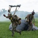 Two military paratroopers have just landed on a grassy field, with their parachute canopy collapsed on the ground behind them. Both personnel are wearing camouflage uniforms, full-face helmets, and tactical harnesses. Smoke is visible in the background, suggesting a military exercise or demonstration.