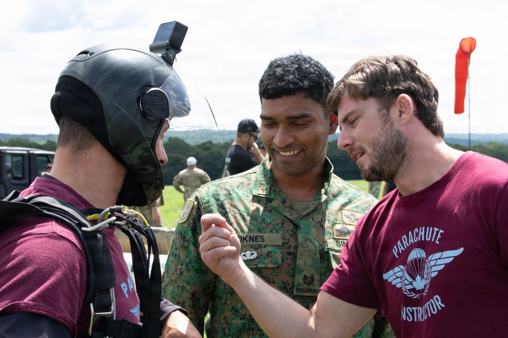 Three individuals are engaged in a close conversation at what appears to be a parachute training or drop zone facility. One person is wearing a jump helmet with a camera mount and parachute harness, while a soldier in Singaporean Armed Forces camouflage uniform and a civilian in a 'Parachute Instructor' maroon t-shirt are interacting with him. The setting includes an open field with a wind sock visible in the background.