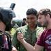 Three individuals are engaged in a close conversation at what appears to be a parachute training or drop zone facility. One person is wearing a jump helmet with a camera mount and parachute harness, while a soldier in Singaporean Armed Forces camouflage uniform and a civilian in a 'Parachute Instructor' maroon t-shirt are interacting with him. The setting includes an open field with a wind sock visible in the background.