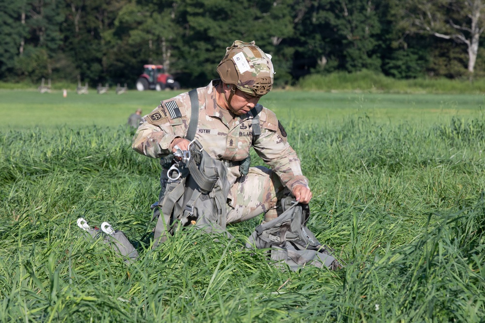 A U.S. Army soldier wearing OCP (Operational Camouflage Pattern) uniform and a tactical helmet is kneeling in a grassy field, gathering and recovering parachute equipment after a jump. The name tape 'HUYNH' is visible on the soldier's chest, along with rank insignia and unit patches including a U.S. flag patch. A red tractor and additional personnel are visible in the background.
