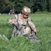 A U.S. Army soldier wearing OCP (Operational Camouflage Pattern) uniform and a tactical helmet is kneeling in a grassy field, gathering and recovering parachute equipment after a jump. The name tape 'HUYNH' is visible on the soldier's chest, along with rank insignia and unit patches including a U.S. flag patch. A red tractor and additional personnel are visible in the background.