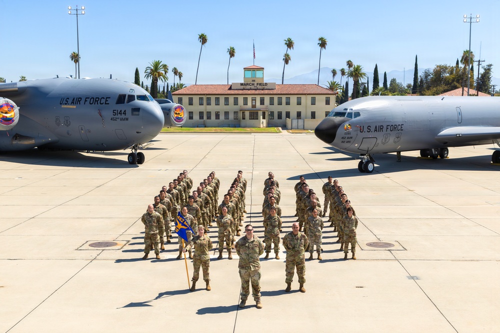 A large group of U.S. Air Force personnel in OCP (Operational Camouflage Pattern) uniforms are posed in formation on a flight line at March Field (March Air Reserve Base), California. Two U.S. Air Force aircraft flank the formation, with the historic March Field building visible in the background. A guidon bearer is visible in the front of the formation, suggesting this is an official unit photograph.
