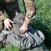 A person wearing military multicam/OCP pattern camouflage uniform is kneeling in tall green grass, handling what appears to be a parachute or large gray fabric equipment bag. The individual is manipulating the gear with both hands, with a red handle or component visible on the equipment. A patch is visible on the left shoulder of the uniform.
