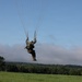 A military paratrooper is captured mid-descent, suspended by parachute suspension lines just above a green grassy field. The individual is wearing military combat uniform and a helmet, with legs raised in a landing preparation posture. The background features a treeline, low clouds, and a clear blue sky.