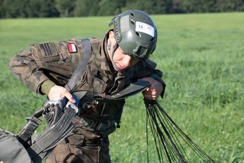 A military paratrooper wearing Polish Army camouflage uniform is gathering parachute lines after a landing on a grassy field. The soldier wears a tactical helmet marked with the number '18' and '18.3', suggesting a competition or exercise event. The individual is actively managing parachute suspension lines and harness equipment post-landing.