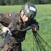 A military paratrooper wearing Polish Army camouflage uniform is gathering parachute lines after a landing on a grassy field. The soldier wears a tactical helmet marked with the number '18' and '18.3', suggesting a competition or exercise event. The individual is actively managing parachute suspension lines and harness equipment post-landing.