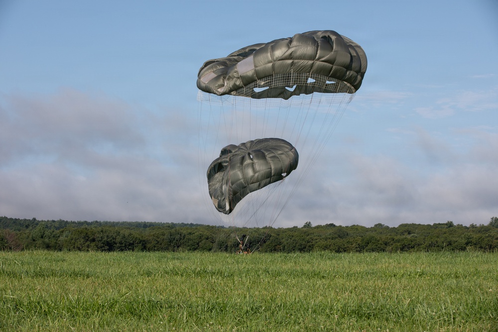 Two olive-drab military parachutes are captured mid-descent over a green grassy field, with a treeline visible in the background. The lower parachute appears partially collapsed or deflating, suggesting the jumper has recently landed or is very close to the ground. A small figure is barely visible at the base of the suspension lines near ground level.