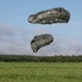 Two olive-drab military parachutes are captured mid-descent over a green grassy field, with a treeline visible in the background. The lower parachute appears partially collapsed or deflating, suggesting the jumper has recently landed or is very close to the ground. A small figure is barely visible at the base of the suspension lines near ground level.