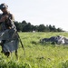 A military paratrooper is standing in a grassy field after a landing, gathering and securing parachute equipment. A deployed parachute canopy lies on the ground behind him, connected by suspension lines. Additional personnel and a vehicle are visible in the background near a tree line.