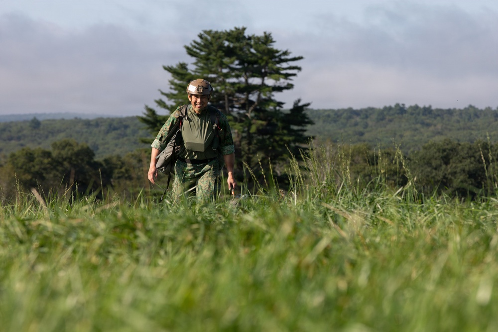 A soldier in military camouflage uniform walks through a tall grass field with a forested hillside in the background. The individual is wearing a helmet with a mounted light, a tactical vest, and carrying a bag or pack. The image appears to be taken during daylight hours in a rural or training environment.