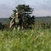 A soldier in military camouflage uniform walks through a tall grass field with a forested hillside in the background. The individual is wearing a helmet with a mounted light, a tactical vest, and carrying a bag or pack. The image appears to be taken during daylight hours in a rural or training environment.