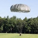 A military paratrooper is landing in an open grassy field using a large olive-drab/sage green round parachute. The jumper appears to be in the final moments of descent, very close to or touching the ground. The setting is a cleared drop zone bordered by dense coniferous trees under a clear blue sky.
