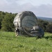 A person in military-style camouflage clothing is managing a large olive-drab/sage green parachute that has landed and is partially inflated on a grassy field. The parachute canopy is billowing and bunched on the ground, with netting visible around its lower sections. The scene appears to be a post-landing parachute recovery operation in a rural, wooded area.