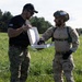 Two individuals in military/tactical attire are standing in a grassy field, sharing what appears to be a Dunkin' Donuts box. The person on the left wears a black t-shirt with a US flag patch, a black cap, and multicam pants, while the person on the right is in full tactical gear including a helmet with the number 9 visible. Both appear to be in good spirits, smiling and interacting in what looks like a training or exercise environment.