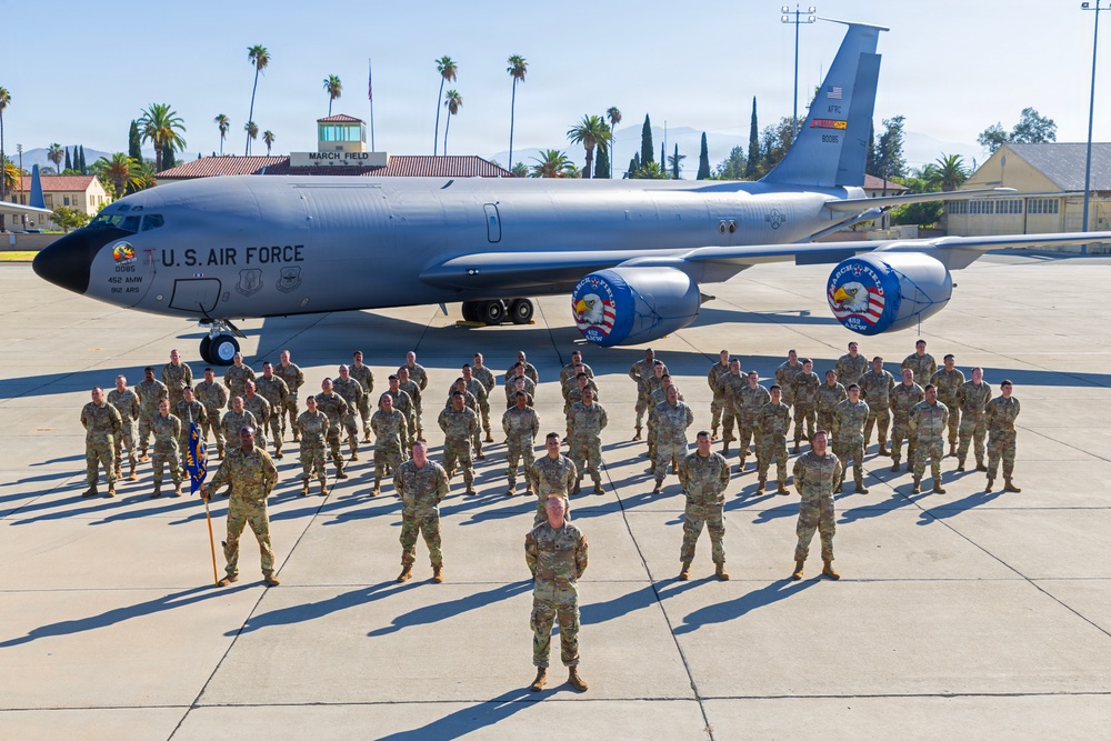A large group of U.S. Air Force personnel in camouflage uniforms are posed in formation on a flight line apron in front of a KC-135 Stratotanker aircraft. The aircraft bears 'U.S. Air Force' markings, unit designations including '452 AMW' and '912 ARS', and decorative nose art featuring eagle emblems with 'March Field' text. The setting is March Air Reserve Base (March Field) in California, identifiable by the control tower sign and surrounding palm trees.