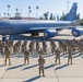 A large group of U.S. Air Force personnel in camouflage uniforms are posed in formation on a flight line apron in front of a KC-135 Stratotanker aircraft. The aircraft bears 'U.S. Air Force' markings, unit designations including '452 AMW' and '912 ARS', and decorative nose art featuring eagle emblems with 'March Field' text. The setting is March Air Reserve Base (March Field) in California, identifiable by the control tower sign and surrounding palm trees.