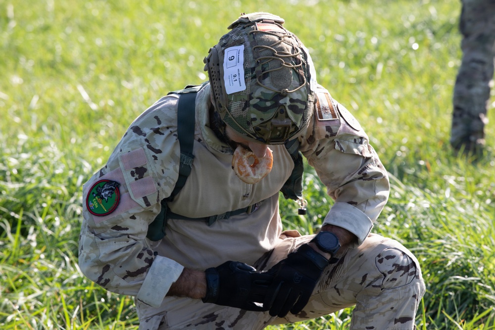 A soldier in desert camouflage (arid pattern) uniform is kneeling on grass with their head bowed down, appearing to eat a donut or pastry without using hands. The soldier is wearing a tactical helmet with a competition number tag '9' attached, along with full combat gear including gloves and a smartwatch. This appears to be part of a military competition or challenge event based on the numbered bib on the helmet.