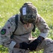 A soldier in desert camouflage (arid pattern) uniform is kneeling on grass with their head bowed down, appearing to eat a donut or pastry without using hands. The soldier is wearing a tactical helmet with a competition number tag '9' attached, along with full combat gear including gloves and a smartwatch. This appears to be part of a military competition or challenge event based on the numbered bib on the helmet.