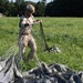 A military paratrooper in desert camouflage uniform and helmet is gathering and recovering a parachute after landing in a green grassy field. The soldier is actively collecting the parachute lines and canopy, which is spread across the ground. Additional personnel and trees are visible in the background.