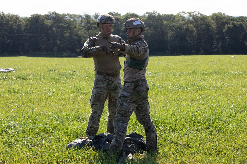 Two military personnel in full camouflage uniforms and tactical helmets are standing in an open grassy field, appearing to assist or check each other's equipment. Dark gear or parachute equipment is visible on the ground at their feet. The setting suggests a post-jump landing zone or field training exercise.