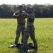 Two military personnel in full camouflage uniforms and tactical helmets are standing in an open grassy field, appearing to assist or check each other's equipment. Dark gear or parachute equipment is visible on the ground at their feet. The setting suggests a post-jump landing zone or field training exercise.