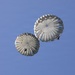 Two military-style parachutes are deployed in a clear blue sky, each carrying a single jumper suspended beneath them. The parachutes are round canopy type, consistent with military static-line parachutes, and appear to be in a descent phase. The image is taken from a low angle looking upward, providing a clear view of both canopies.