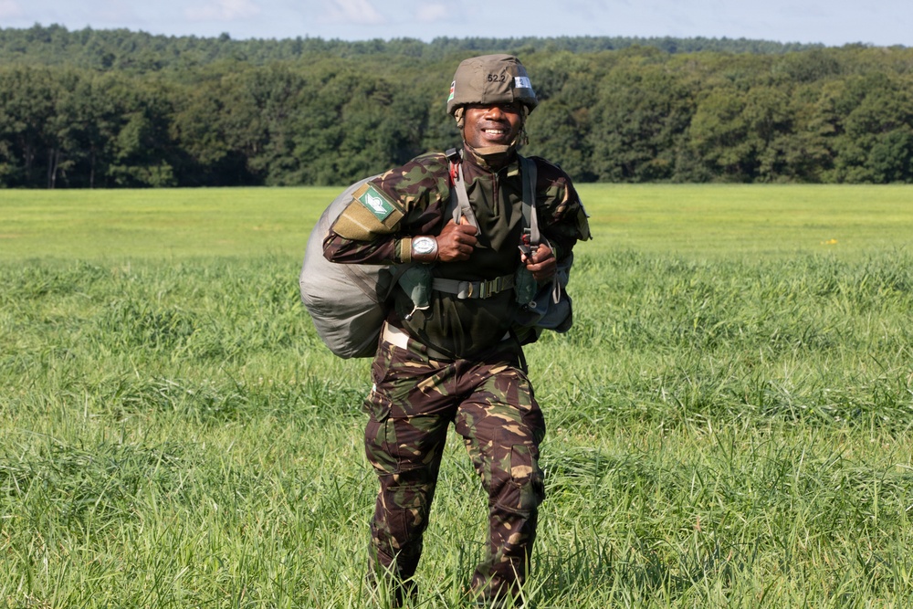 A smiling soldier in camouflage uniform stands in an open green field after what appears to be a parachute landing, carrying a bundled parachute pack. The soldier is wearing a helmet with a number visible and has shoulder patches on the uniform. A treeline is visible in the background under a partly cloudy sky.