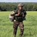 A smiling soldier in camouflage uniform stands in an open green field after what appears to be a parachute landing, carrying a bundled parachute pack. The soldier is wearing a helmet with a number visible and has shoulder patches on the uniform. A treeline is visible in the background under a partly cloudy sky.