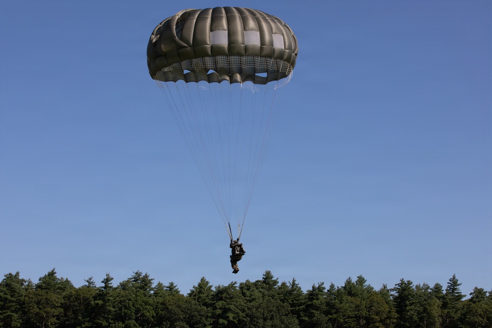 A military paratrooper is descending under a large olive-drab/khaki round parachute against a clear blue sky. The jumper appears to be approaching a landing zone just above a treeline of dense evergreen trees. The parachute is fully deployed and appears to be a standard military static-line round canopy.