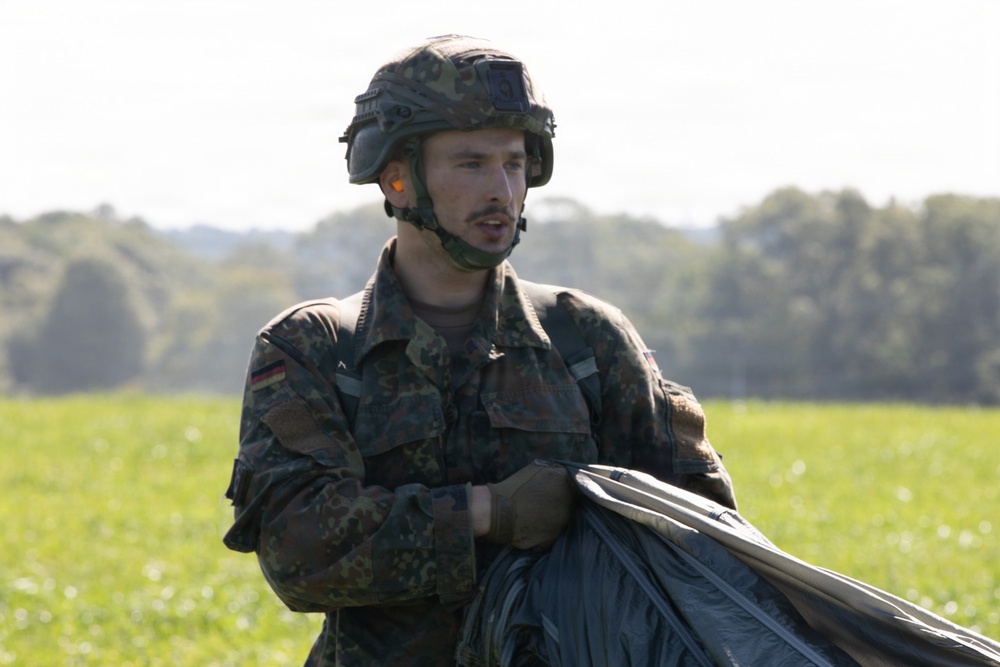A male soldier wearing German Bundeswehr flecktarn camouflage uniform and combat helmet is standing in an open grassy field. He appears to be gathering or handling what looks like a parachute canopy. The background shows green trees and an overcast sky, suggesting an outdoor military exercise or post-jump recovery.
