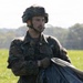 A male soldier wearing German Bundeswehr flecktarn camouflage uniform and combat helmet is standing in an open grassy field. He appears to be gathering or handling what looks like a parachute canopy. The background shows green trees and an overcast sky, suggesting an outdoor military exercise or post-jump recovery.