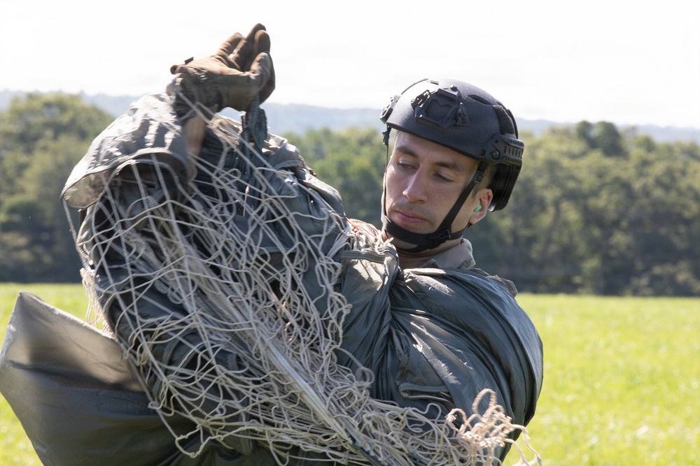 A military personnel is handling what appears to be a parachute canopy and suspension lines in an open grassy field. The individual is wearing tactical gear including a ballistic helmet and gloves, and appears to be gathering or packing the parachute equipment after a jump. The background shows a treeline and open green field under overcast daylight conditions.