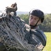 A military personnel is handling what appears to be a parachute canopy and suspension lines in an open grassy field. The individual is wearing tactical gear including a ballistic helmet and gloves, and appears to be gathering or packing the parachute equipment after a jump. The background shows a treeline and open green field under overcast daylight conditions.