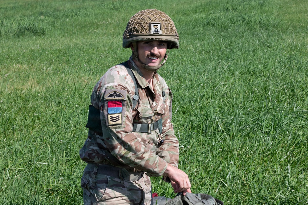 A smiling male soldier in British MTP (Multi-Terrain Pattern) camouflage uniform is kneeling or crouching in a green grassy field. He wears a helmet with a camouflage net cover and carries visible rank insignia and unit patches on his sleeve. His relaxed expression and posture suggest a pause during a training exercise or military event.