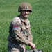 A smiling male soldier in British MTP (Multi-Terrain Pattern) camouflage uniform is kneeling or crouching in a green grassy field. He wears a helmet with a camouflage net cover and carries visible rank insignia and unit patches on his sleeve. His relaxed expression and posture suggest a pause during a training exercise or military event.
