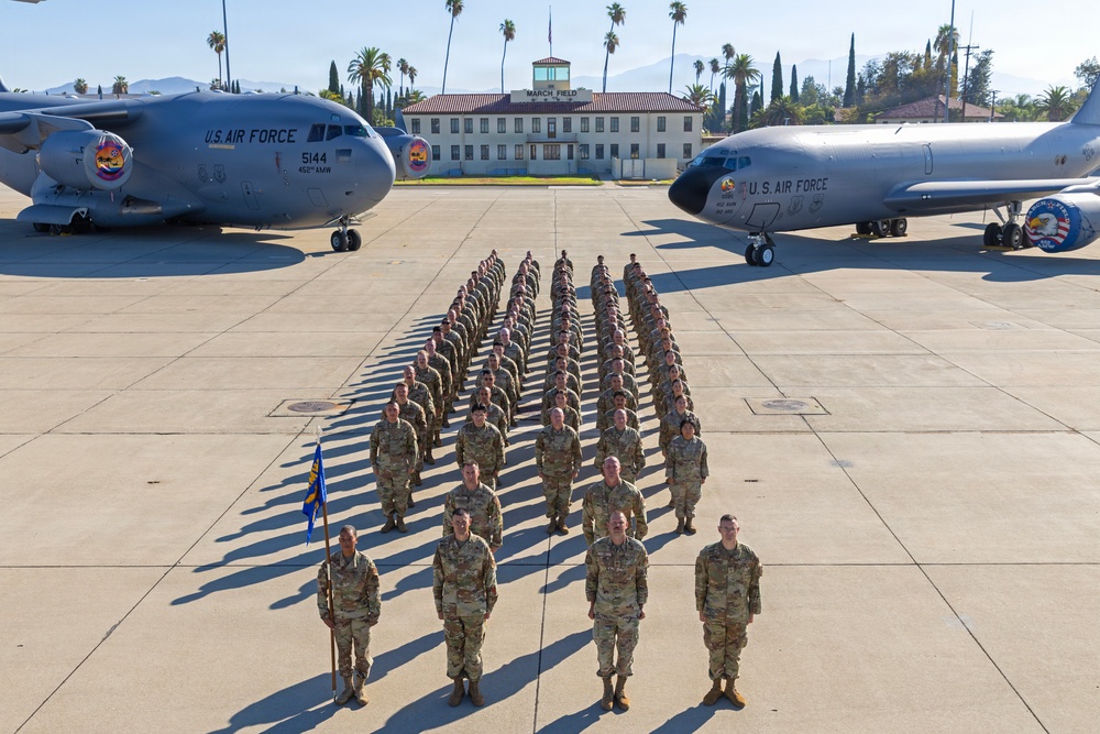 A formal military formation photograph taken on the flight line at March Field (March Air Reserve Base), California. Personnel in OCP (Operational Camouflage Pattern) uniforms are arranged in precise rows between two U.S. Air Force aircraft. A guidon bearer stands at the front-left of the formation alongside what appear to be senior officers or NCOs.