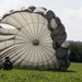 A large military parachute has landed and is still partially inflated on a green grassy field. At least three personnel in military uniforms are visible around the canopy, appearing to manage or recover the parachute after a jump. The setting is a rural open field with tree lines and hills visible in the background under a partly cloudy sky.