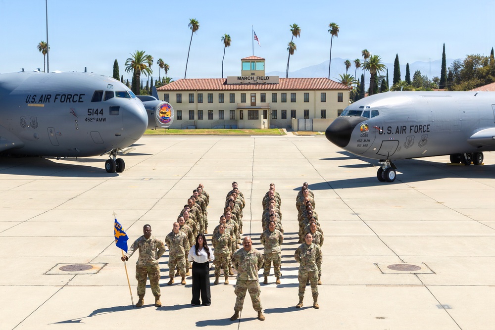 A group of U.S. Air Force personnel pose for a formal unit photograph on the flight line at March Air Reserve Base (March Field), California. Two large military transport aircraft flank the formation, with the historic March Field headquarters building visible in the background. One civilian woman in white and black attire stands among the uniformed service members, and a unit guidon flag is being held on the left side of the formation.
