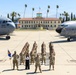 A group of U.S. Air Force personnel pose for a formal unit photograph on the flight line at March Air Reserve Base (March Field), California. Two large military transport aircraft flank the formation, with the historic March Field headquarters building visible in the background. One civilian woman in white and black attire stands among the uniformed service members, and a unit guidon flag is being held on the left side of the formation.