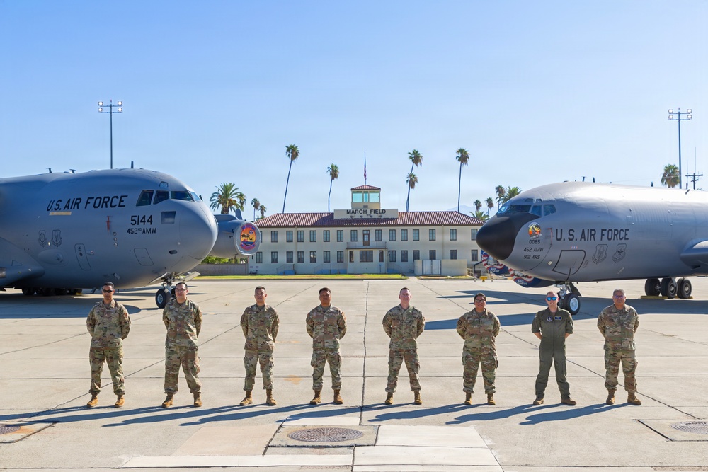 Eight U.S. Air Force personnel in OCP (Operational Camouflage Pattern) uniforms are posed on a flight line at March Air Reserve Base (March Field) in California. Two large U.S. Air Force aircraft are positioned on either side of the group, with the historic March Field headquarters building visible in the background. The aircraft markings indicate assignment to the 452nd Air Mobility Wing.