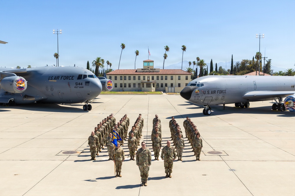 A formal military unit formation photograph taken on the flight line at March Field Air Reserve Base, with personnel in OCP (Operational Camouflage Pattern) uniforms arranged in a V-shaped formation. Two U.S. Air Force aircraft flank the formation — a C-17 Globemaster III (tail number 5144, 452nd AMW) on the left and what appears to be a KC-135 Stratotanker on the right. The historic March Field headquarters building is visible in the background with an American flag flying above it.