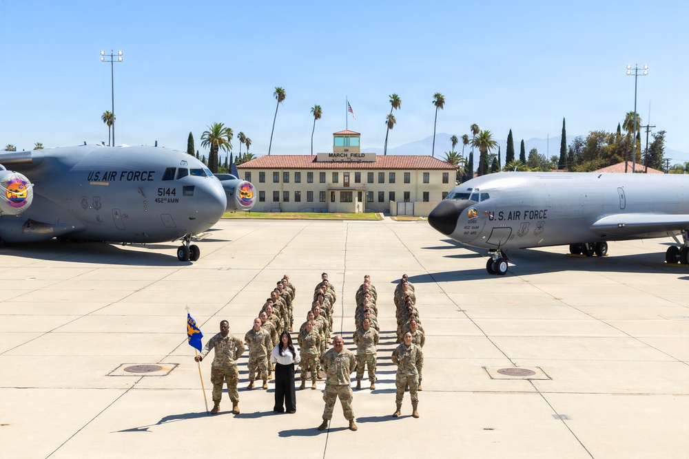A group of U.S. Air Force personnel are posed in formation on the flight line at March Field (March Air Reserve Base) in California, flanked by two military aircraft. One civilian woman in white and black attire stands among the uniformed service members, with a unit guidon flag visible. The historic March Field headquarters building is visible in the background, along with palm trees and mountains.