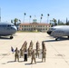 A group of U.S. Air Force personnel are posed in formation on the flight line at March Field (March Air Reserve Base) in California, flanked by two military aircraft. One civilian woman in white and black attire stands among the uniformed service members, with a unit guidon flag visible. The historic March Field headquarters building is visible in the background, along with palm trees and mountains.