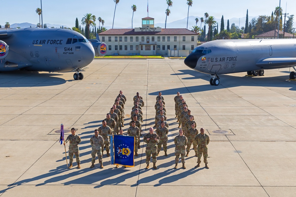 A group of U.S. Air Force personnel in OCP (Operational Camouflage Pattern) uniforms are assembled in formation on the flight line at March Field (March Air Reserve Base) in California. They are flanked by two U.S. Air Force aircraft — a C-17 Globemaster III (tail number 5144, 452nd AMW) on the left and what appears to be a KC-135 Stratotanker on the right. A blue unit guidon/flag is prominently displayed at the front of the formation.