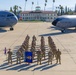 A group of U.S. Air Force personnel in OCP (Operational Camouflage Pattern) uniforms are assembled in formation on the flight line at March Field (March Air Reserve Base) in California. They are flanked by two U.S. Air Force aircraft — a C-17 Globemaster III (tail number 5144, 452nd AMW) on the left and what appears to be a KC-135 Stratotanker on the right. A blue unit guidon/flag is prominently displayed at the front of the formation.