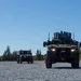 Two military ground vehicles are traveling along a gravel surface marked with orange traffic cones, suggesting a controlled test or demonstration environment. The lead vehicle is a larger, heavily equipped tactical wheeled vehicle with roof-mounted systems and active headlights, while the trailing vehicle appears smaller and unmanned. The setting includes a clear blue sky and tree line in the background, consistent with a military training or evaluation area.