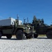 Two military tactical vehicles, appearing to be Joint Light Tactical Vehicles (JLTVs) in olive drab green, are parked on a gravel surface in what appears to be a coastal or tropical environment. The vehicles are heavily equipped with communications and electronic warfare systems, including multiple antennas, sensor arrays, and what appears to be a large equipment shelter or payload box mounted on the rear vehicle. The setting suggests a military exercise or demonstration environment.