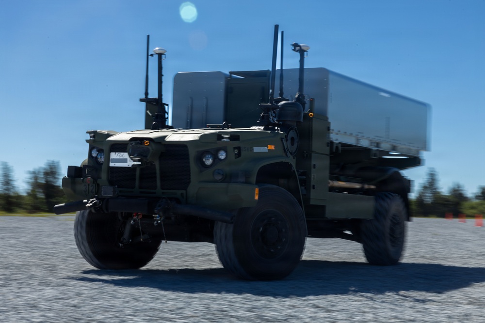 A military tactical wheeled vehicle, appearing to be an autonomous or semi-autonomous variant of a FMTV/LMTV-class truck, is moving across a gravel surface. The vehicle is painted in olive drab/military green and is equipped with multiple sensors, antennas, and what appears to be a LiDAR unit, suggesting unmanned ground vehicle (UGV) capability. The background shows a clear blue sky and tree line, consistent with a military test or demonstration environment.