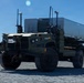 A military tactical wheeled vehicle, appearing to be an autonomous or semi-autonomous variant of a FMTV/LMTV-class truck, is moving across a gravel surface. The vehicle is painted in olive drab/military green and is equipped with multiple sensors, antennas, and what appears to be a LiDAR unit, suggesting unmanned ground vehicle (UGV) capability. The background shows a clear blue sky and tree line, consistent with a military test or demonstration environment.